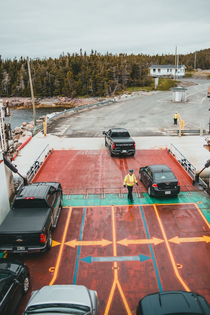 mobile-01 Trucks and cars exit a ferry, guided by workers at a dock in a rural setting.
